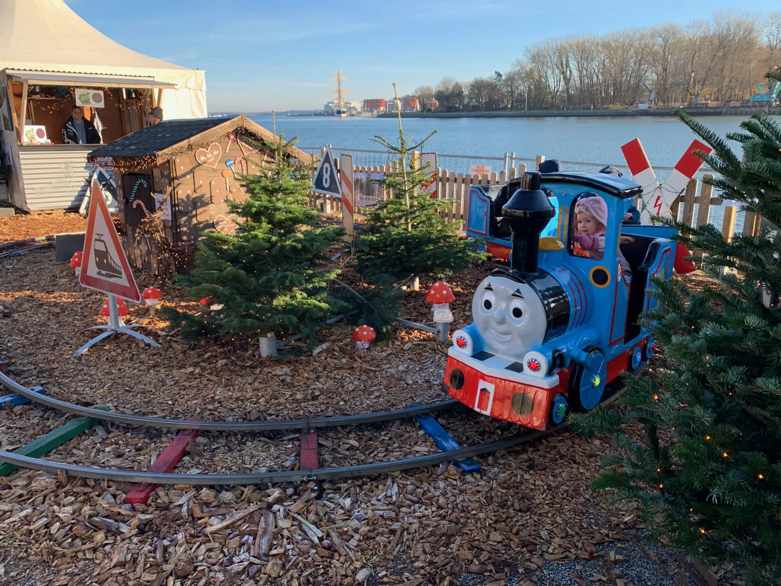 Kleine Kindereisenbahn zwischen Tannenbäumen auf dem Weihnachtsmarkt am Kreuzfahrtterminal in Travemünde, dahinter die Trave